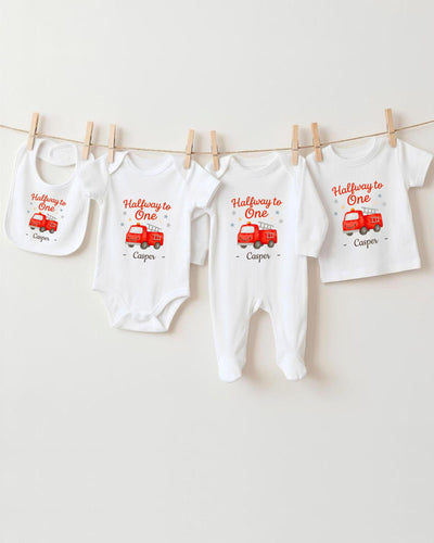 A collection of white baby clothes, including onesies and bibs, with red illustrations of fire trucks and the text "Halfway to One" printed on them.
