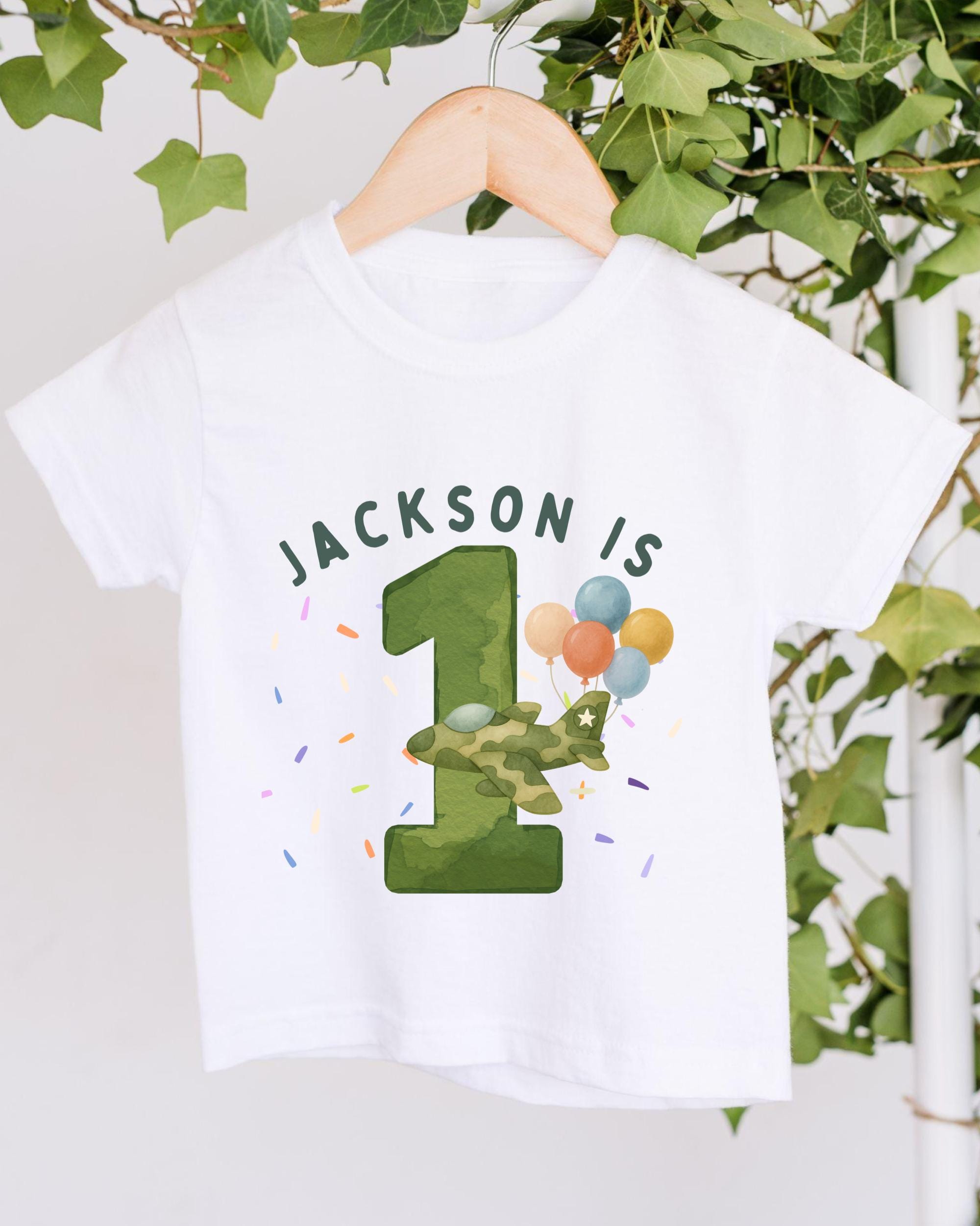 A white t-shirt with a graphic design featuring a green number one, balloons, and the text "JACKSON IS 1" printed on it.