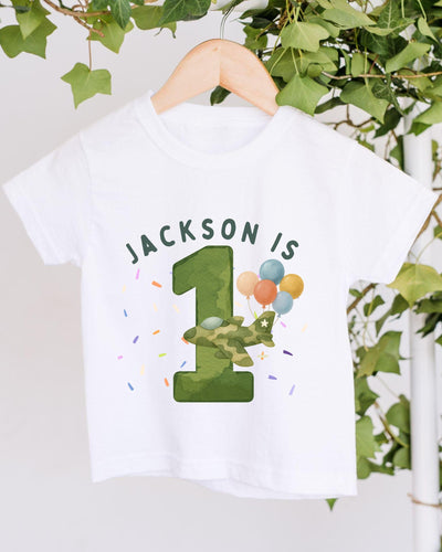 A white t-shirt with a graphic design featuring a green number one, balloons, and the text "JACKSON IS 1" printed on it.