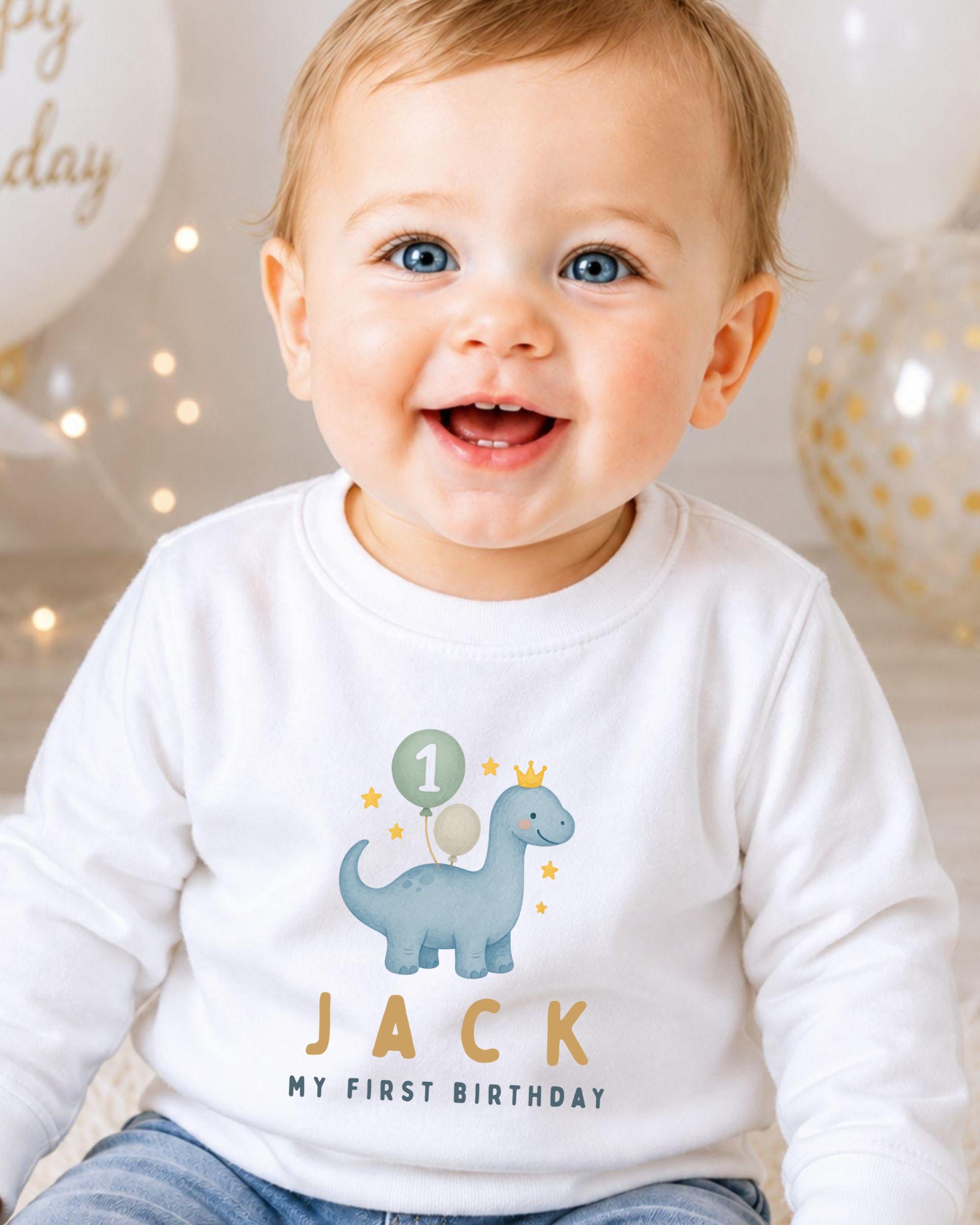 A smiling baby boy wearing a white shirt with a dinosaur and the text "Jack" and "My first birthday" printed on it.