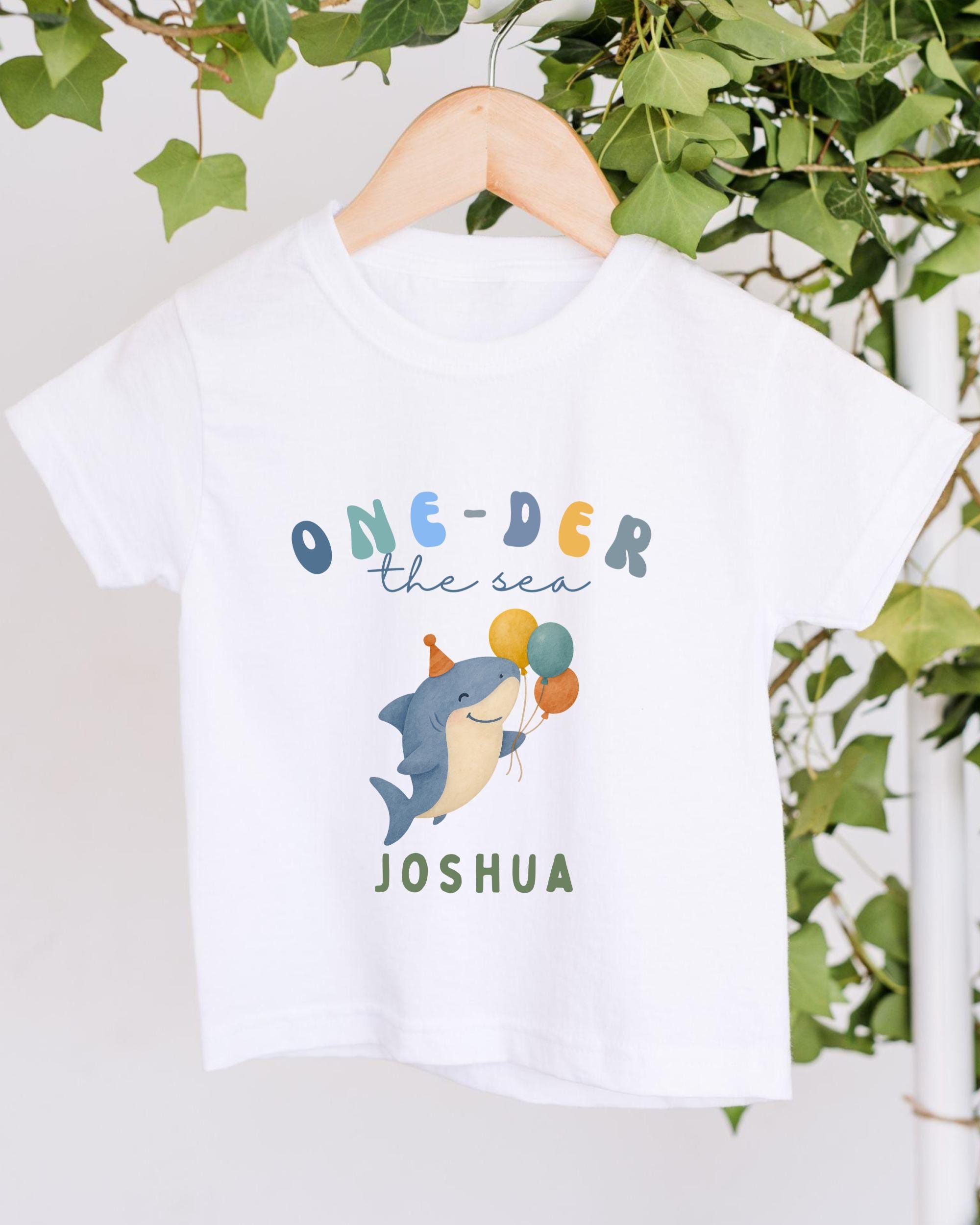 A white t-shirt with a graphic design featuring a cartoon shark holding balloons, along with the text "One-der the sea Joshua" printed on it.