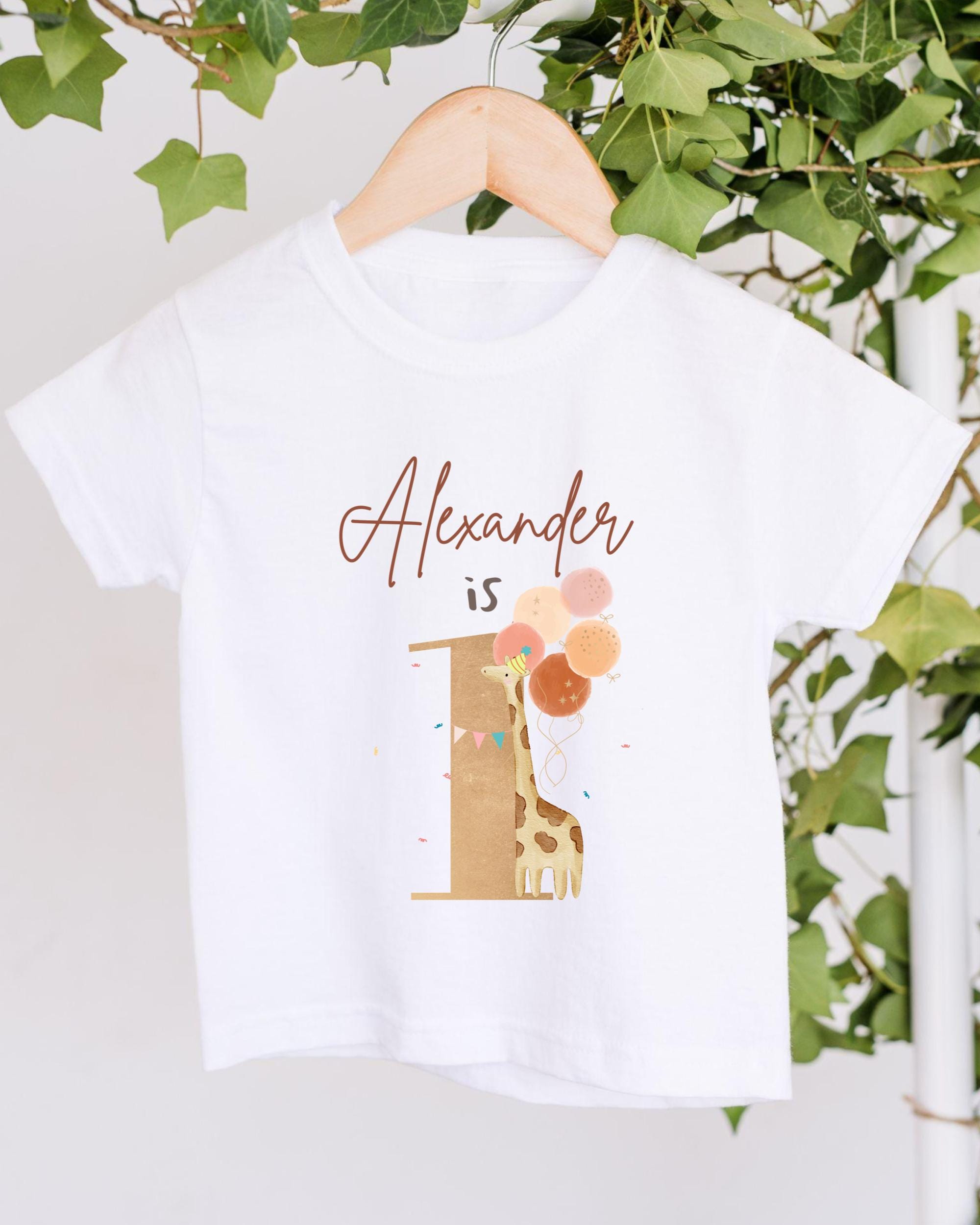 A white t-shirt with a giraffe and balloons printed on it, hanging on a wooden hanger.