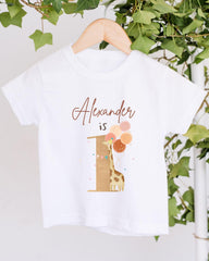 A white t-shirt with a giraffe and balloons printed on it, hanging on a wooden hanger.