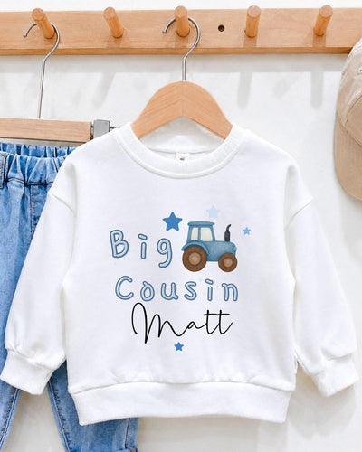 A white sweatshirt with a tractor design and the text "Big Cousin Matt" printed on it, hanging on a wooden hanger.