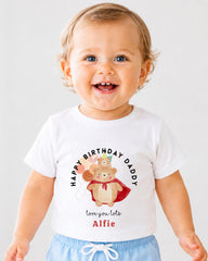 A smiling baby boy wearing a white t-shirt with a birthday message and a cartoon bear graphic.