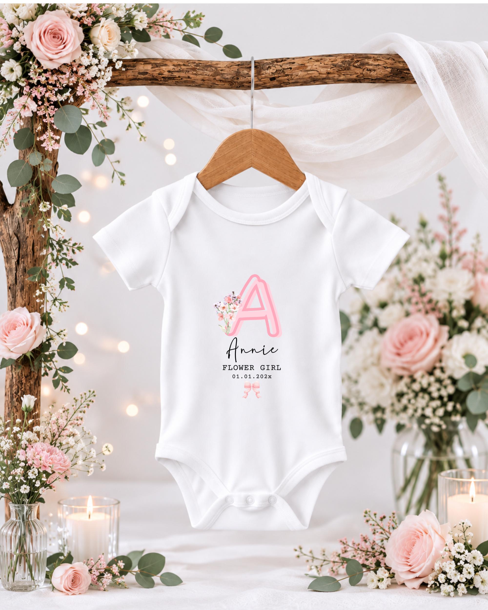 A white baby onesie with a pink letter "A" and the text "Alicie Flower Girl" is hanging on a wooden hanger, surrounded by pink and white flowers, candles, and greenery.