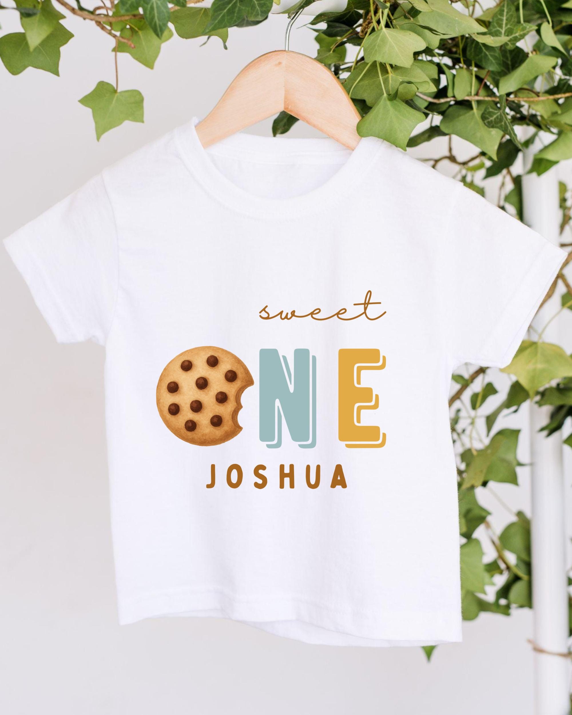 A white t-shirt with a graphic design featuring a cookie and the text "Sweet One Joshua" printed on it.