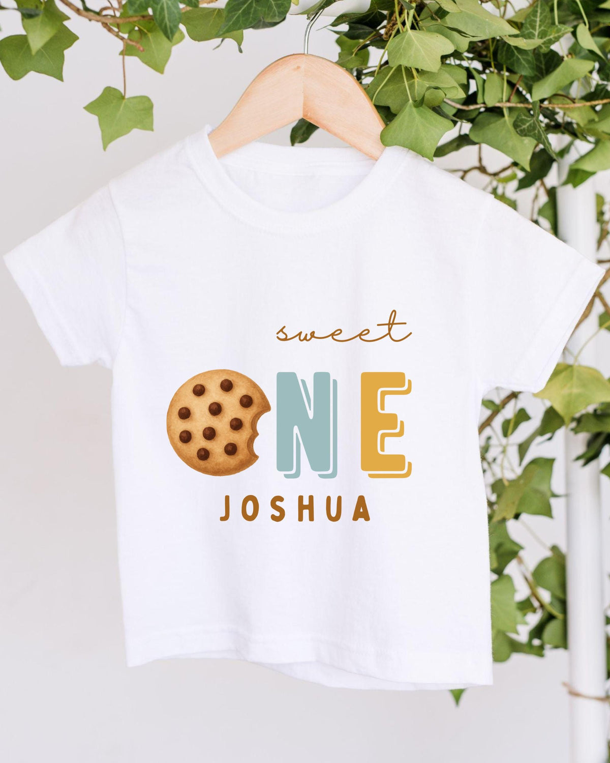 A white t-shirt with a graphic design featuring a cookie and the text "Sweet One Joshua" printed on it.