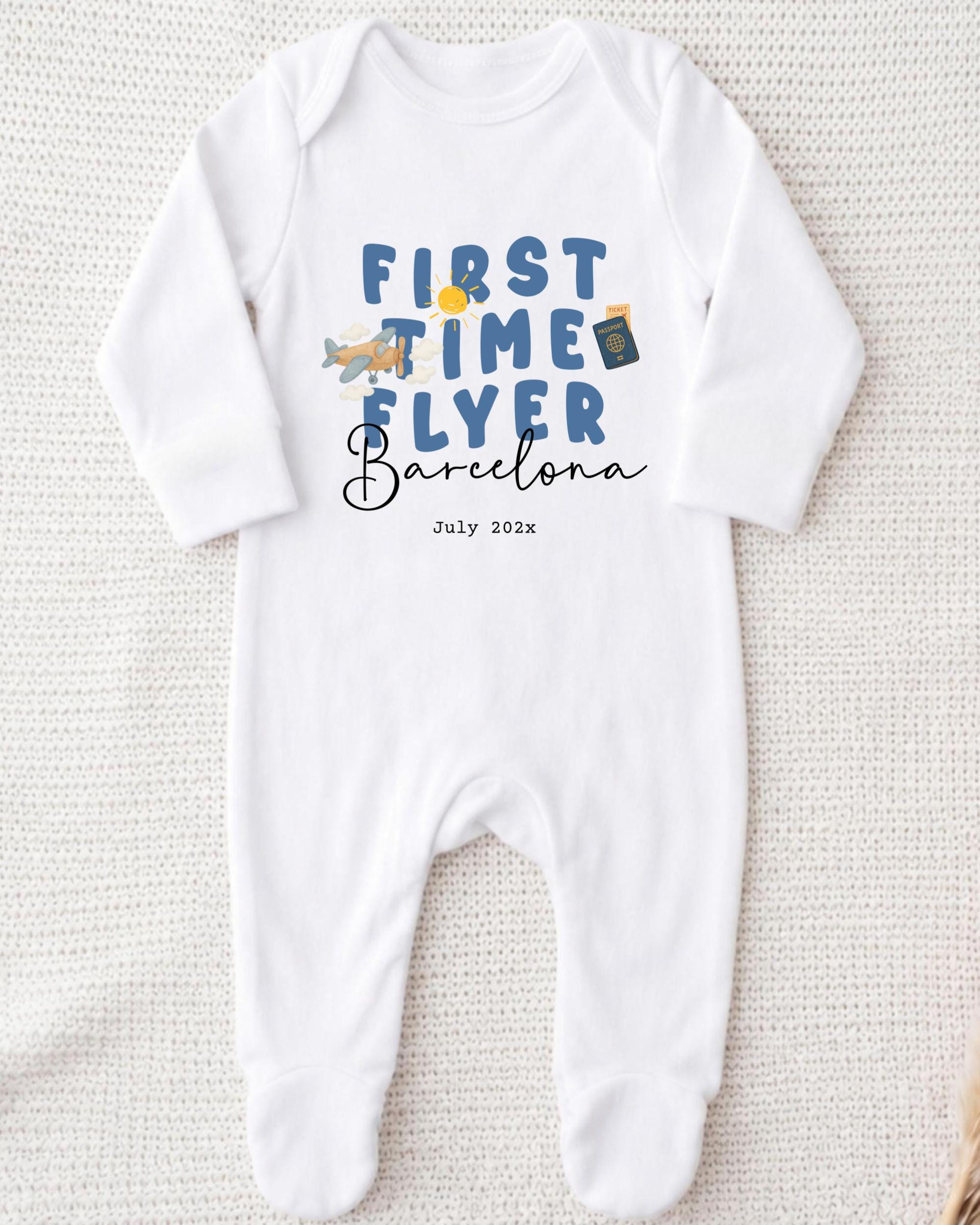A white onesie with the text "FIRST TIME FLYER Barcelona" printed on it, along with a small airplane and a suitcase.