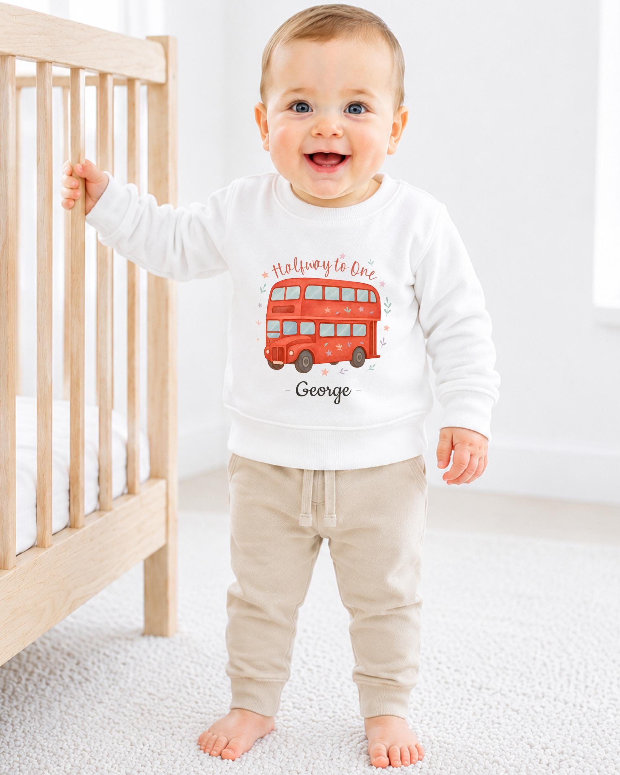 A smiling baby boy wearing a white sweatshirt with a red double-decker bus design and the name 'George' printed on it.