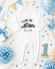 A white onesie with a race car design, surrounded by birthday decorations, confetti, and a blue balloon with the text "Happy Birthday".