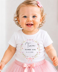 A smiling baby girl wearing a white t-shirt with a floral design and a pink bow in her hair.