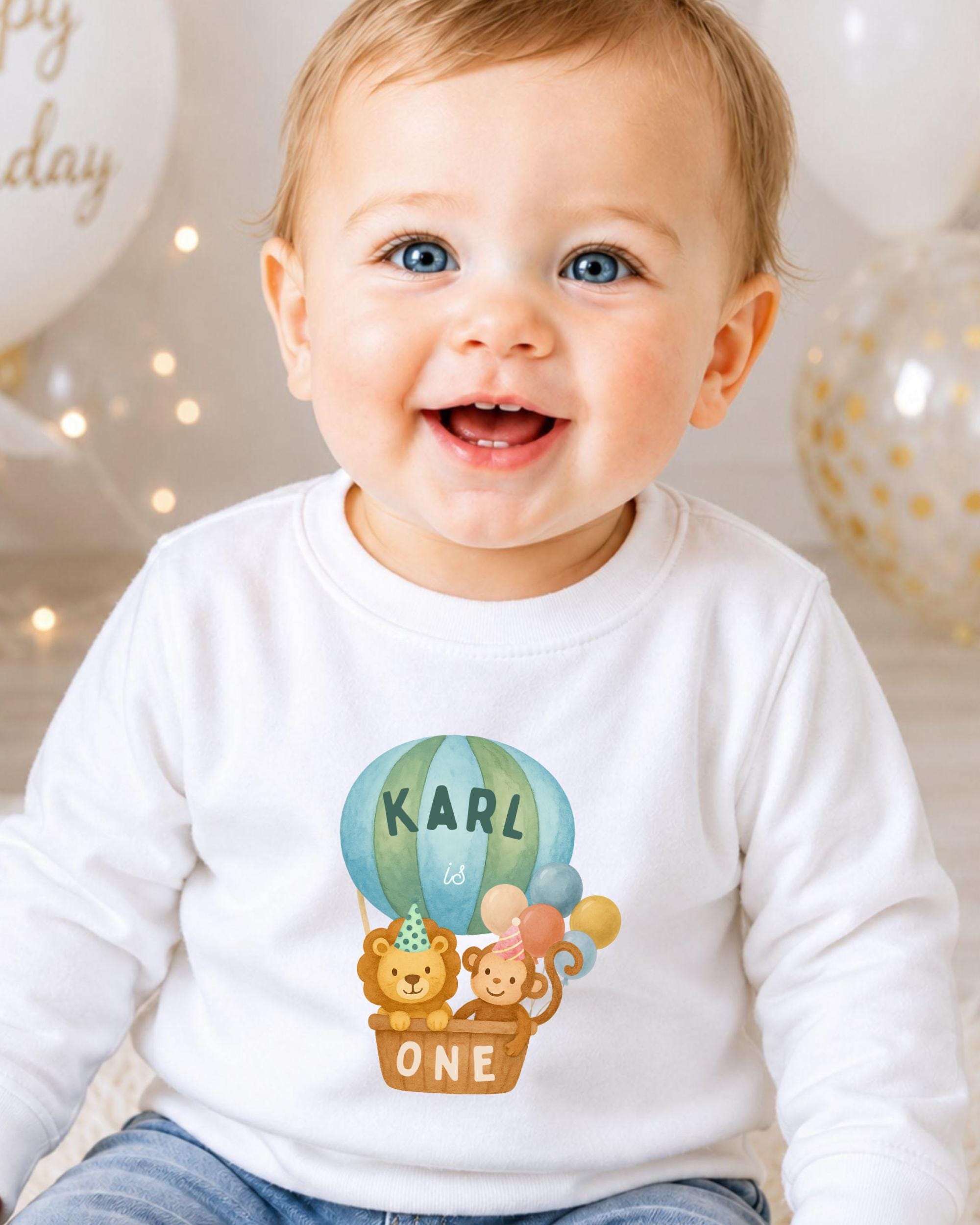 A smiling baby boy wearing a white shirt with a cartoon image of a hot air balloon and animals, including a lion and a monkey, along with the name "KARL" and the number "1" printed on it.