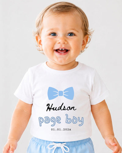 A smiling baby boy wearing a white t-shirt with a blue bow tie and the text "Hudson Page Boy 01.01.2023" printed on it.