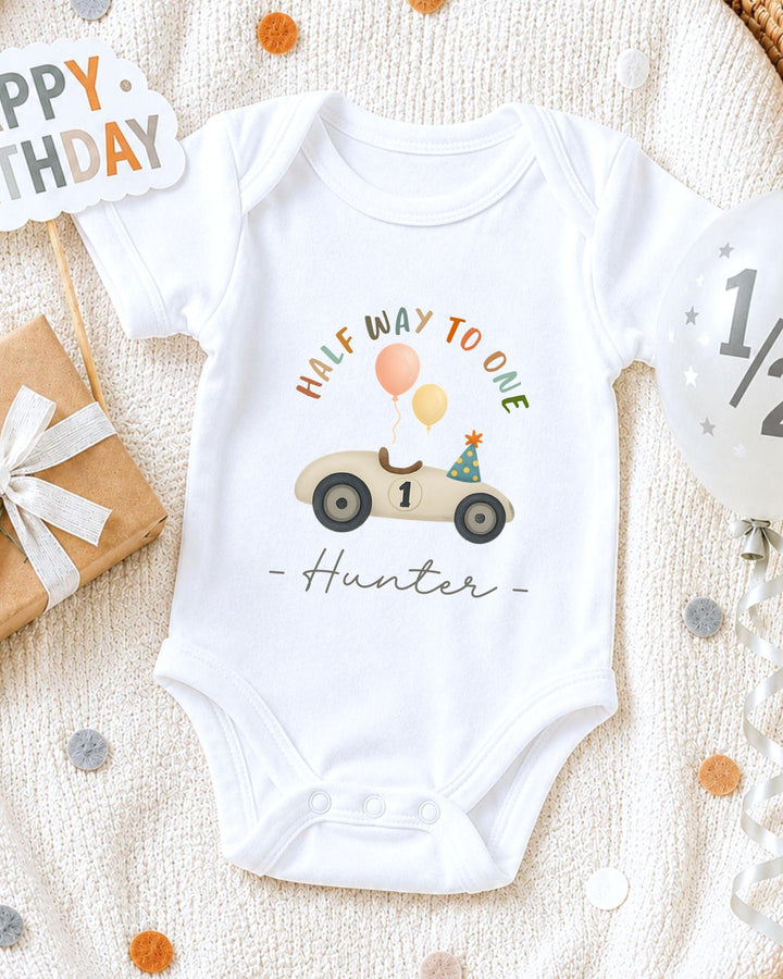 A white onesie with a cartoon car and balloons, along with a "Happy Birthday Hunter" message, is displayed on a bed with confetti.