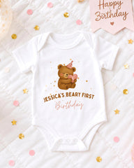 A white onesie with a teddy bear and a birthday message is displayed on a bed with confetti and a birthday card.