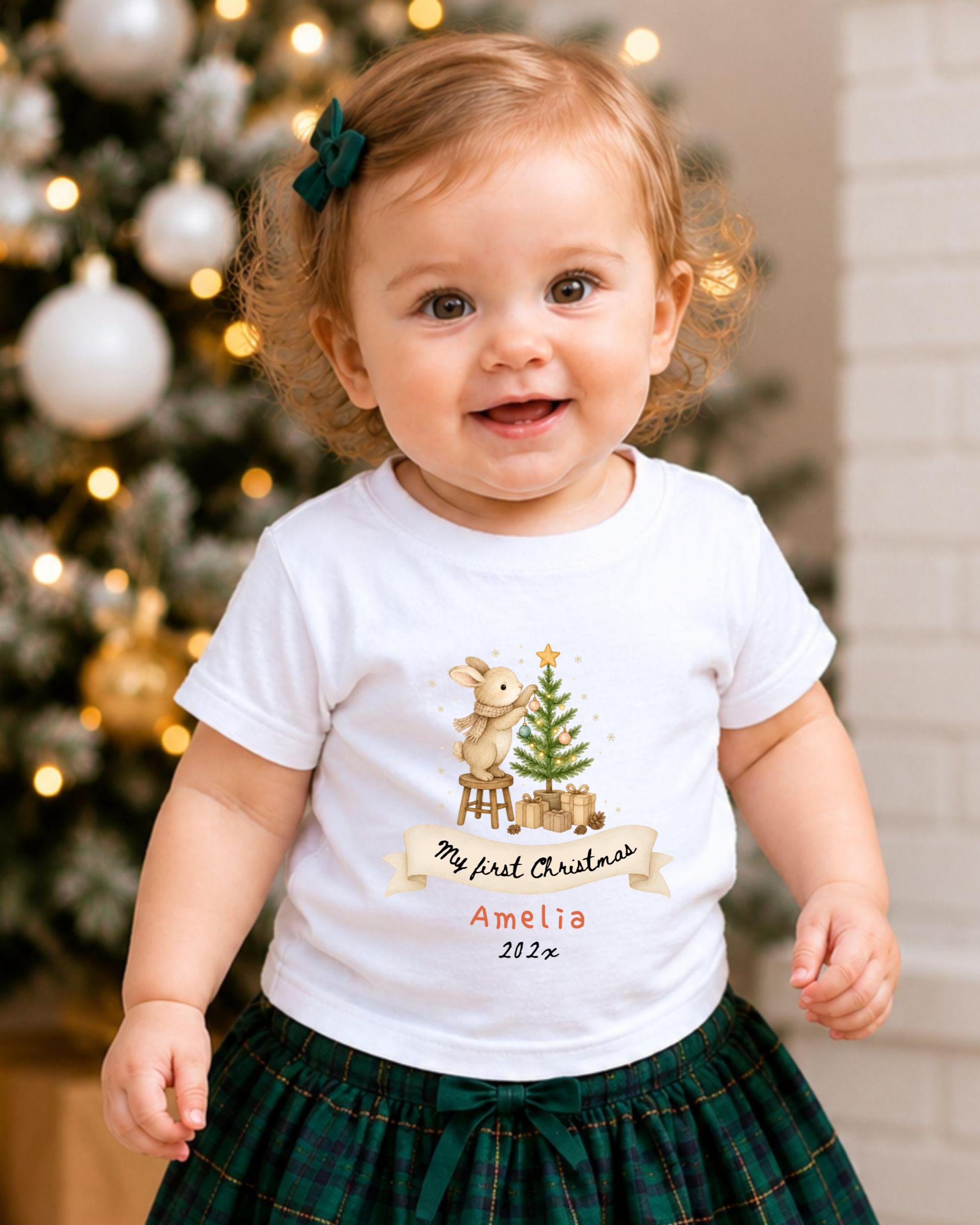 A smiling baby girl wearing a white t-shirt with a Christmas-themed design and a ribbon that says "My first Christmas" and "Amelia 2020".