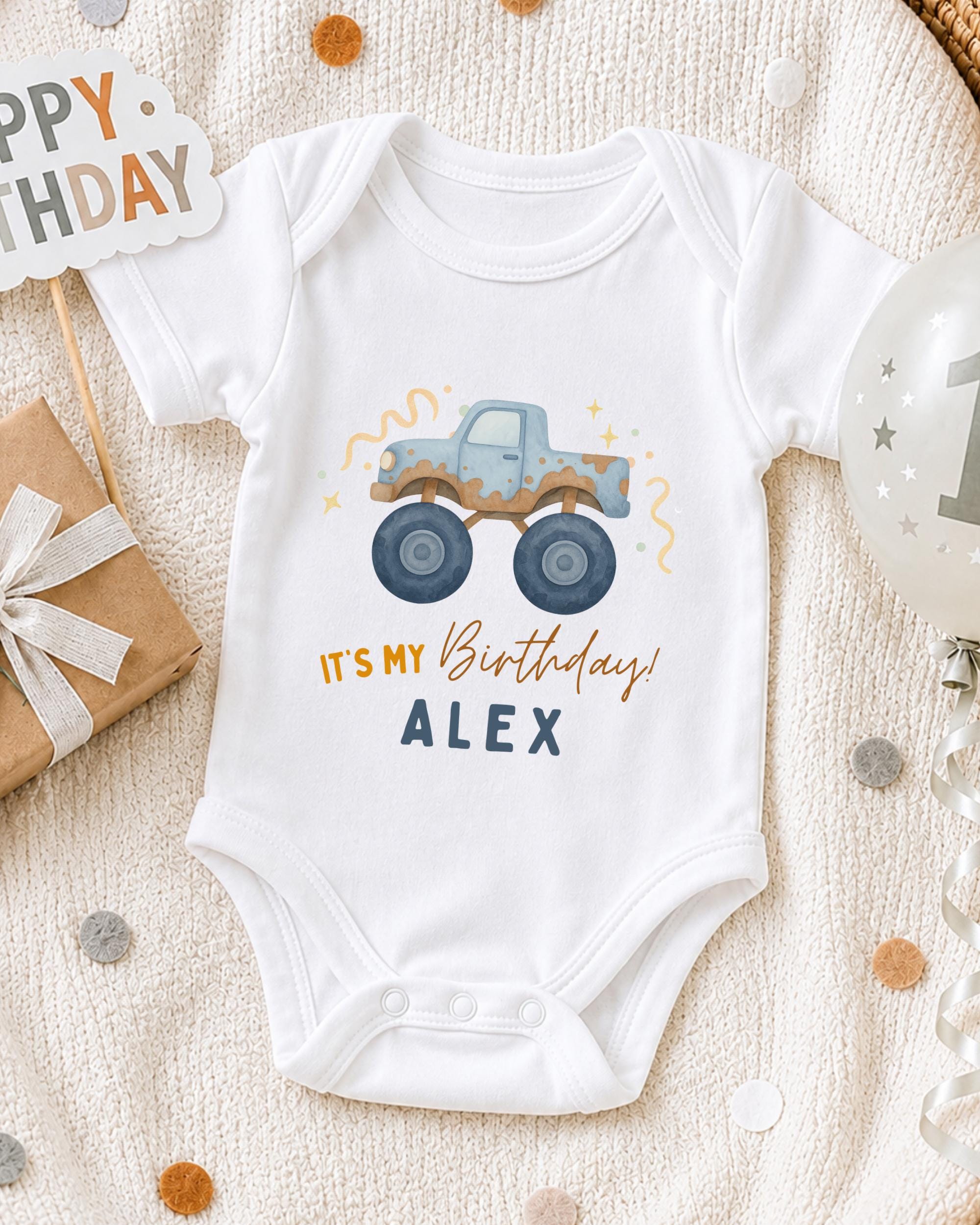 A white onesie with a truck graphic and the text "It's my birthday! ALEX" printed on it, along with a "Happy Birthday" sign and a balloon with the number "1" on it, all placed on a white blanket with confetti.