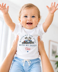 A smiling baby wearing a white t-shirt with a racing car design and the text "Watching the race with my daddy" is being held up by someone's arms.