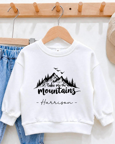 A white sweatshirt with a mountain landscape design and the text "Take me the mountains" printed on it, hanging on a wooden hanger.