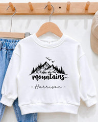 A white sweatshirt with a mountain landscape design and the text "Take me the mountains" printed on it, hanging on a wooden hanger.