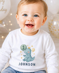 A smiling baby boy wearing a white shirt with a dinosaur graphic and the name "Johnson" printed on it.