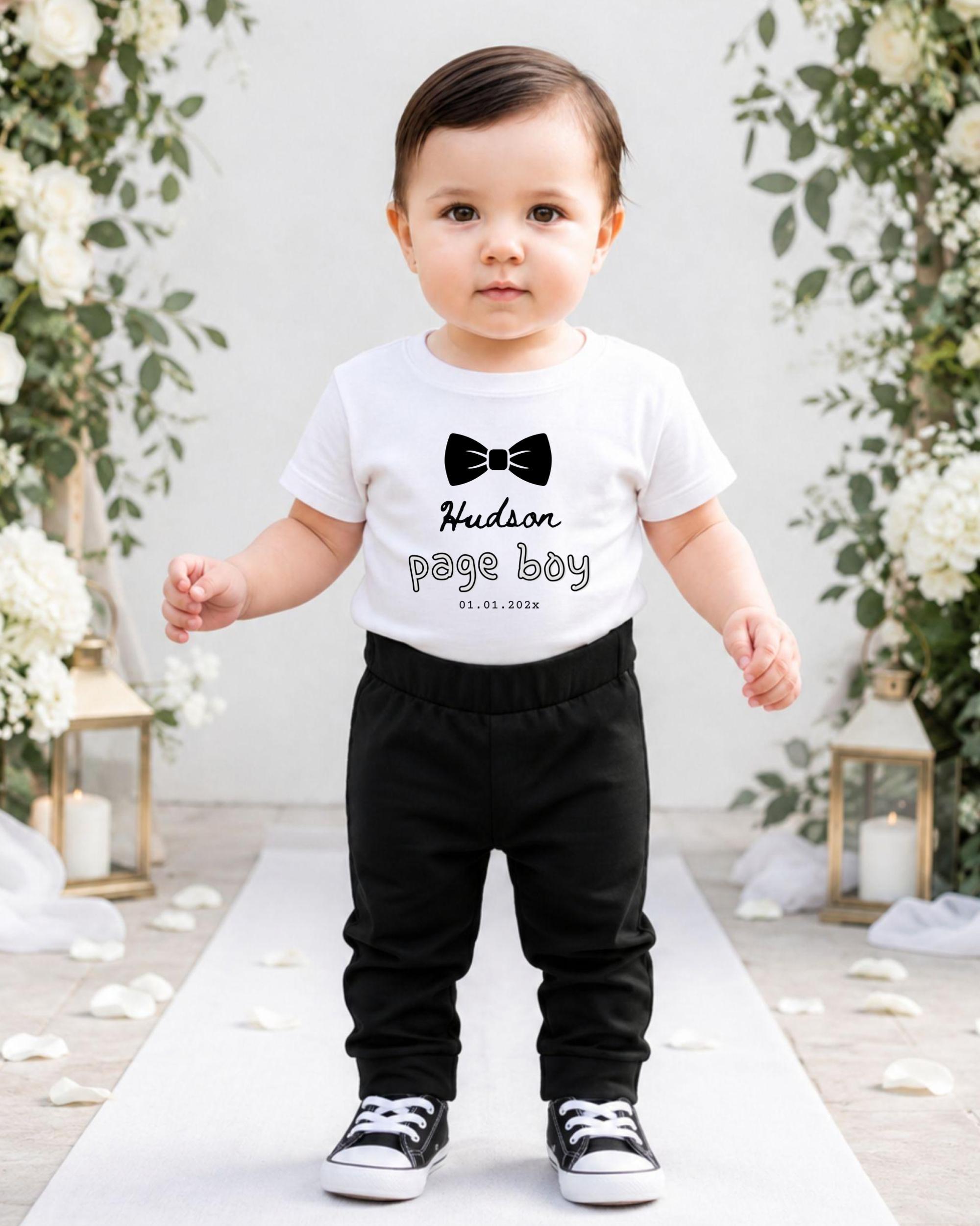 A young child wearing a white t-shirt with a bow tie and black pants stands on a white carpet in front of a floral archway.