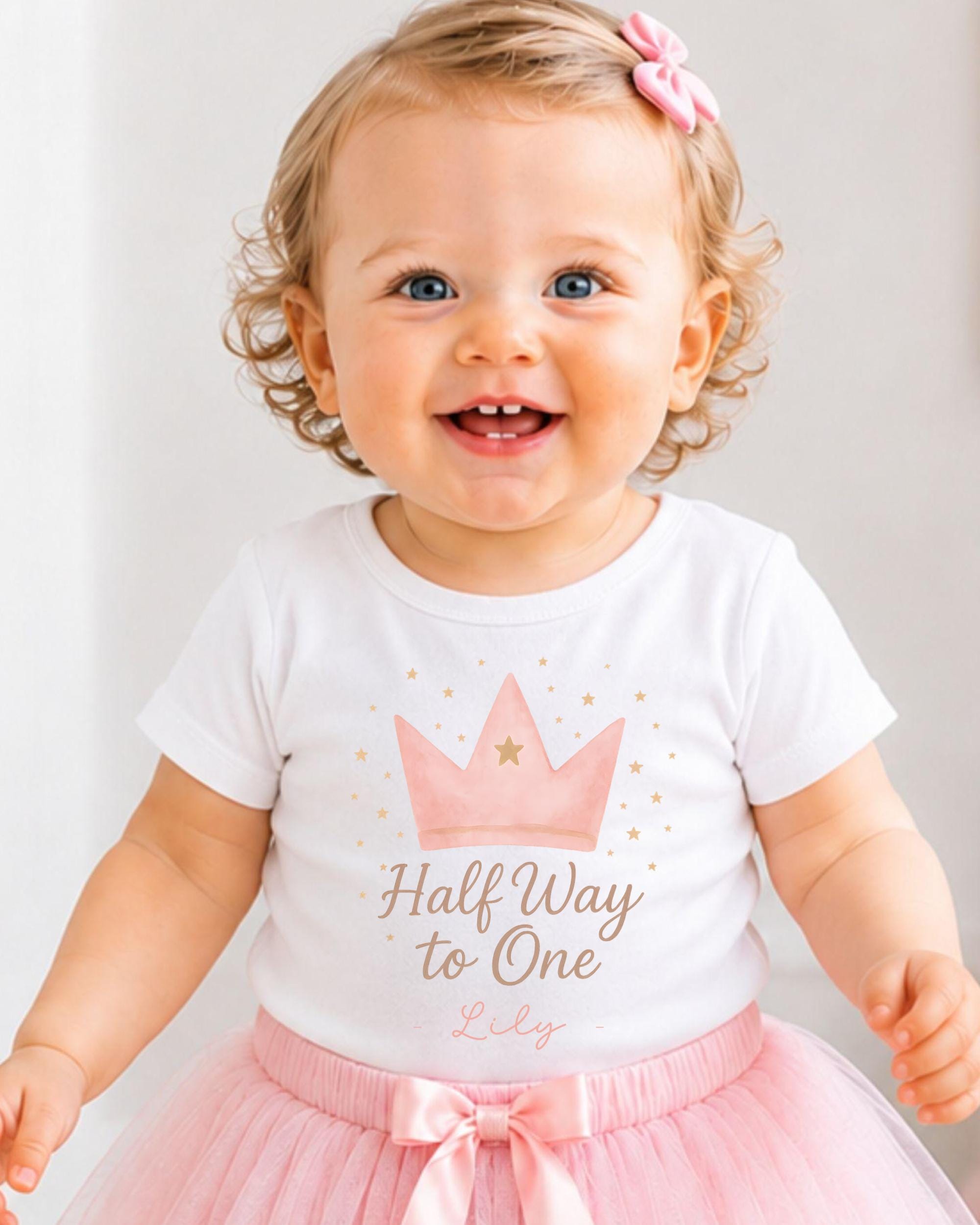 A smiling baby girl wearing a white t-shirt with a crown and stars, and a pink tutu skirt.