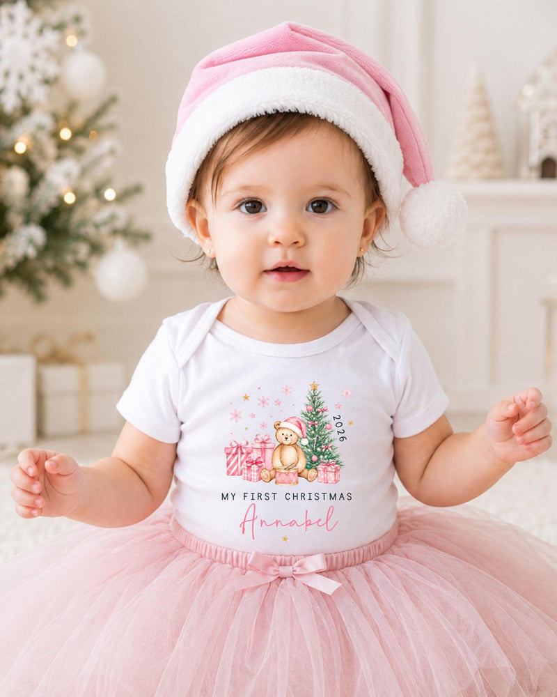 A young girl wearing a pink Santa hat and a white t-shirt with a Christmas-themed design, sitting on a white carpet in front of a Christmas tree.