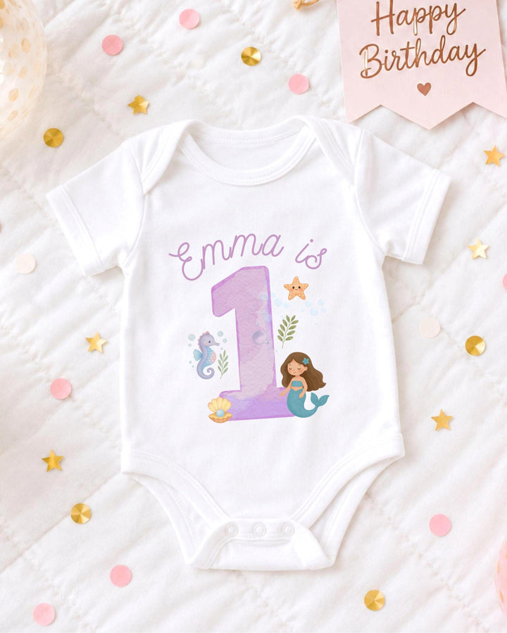 A white bodysuit with a mermaid design and the number 1, surrounded by confetti and a birthday card.