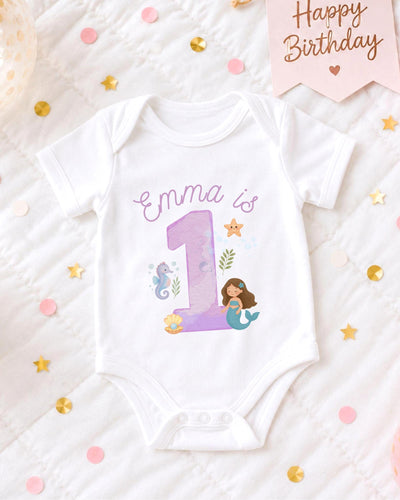 A white bodysuit with a mermaid design and the number 1, surrounded by confetti and a birthday card.