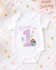 A white onesie with a mermaid design and the number 1, surrounded by confetti and a birthday card.
