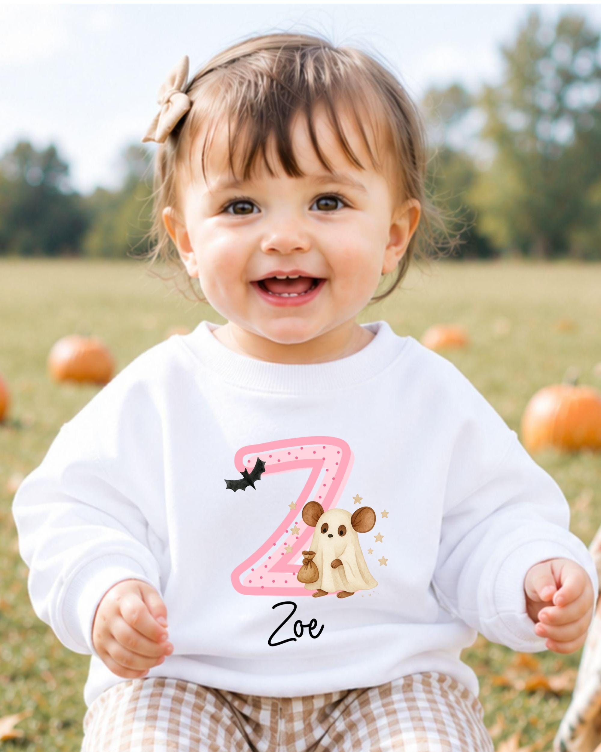 A smiling baby girl wearing a white sweater with a pink letter "Z" and a cartoon character on it, sitting in a field with pumpkins in the background.