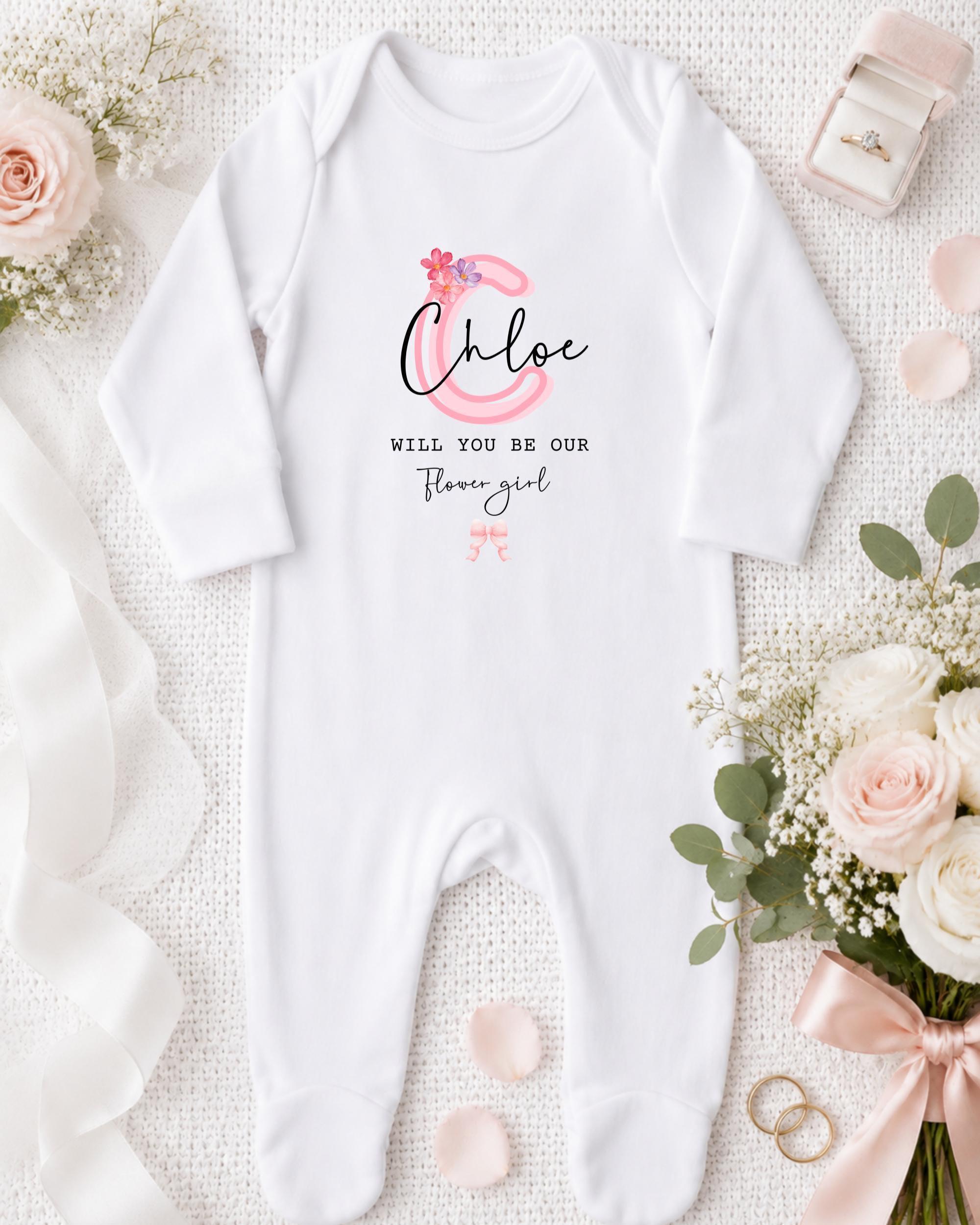 A white onesie with a personalized message, a pink flower, and a pink ribbon, along with a ring box, a bouquet of white flowers, and a white lace ribbon.