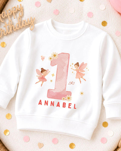 A white sweatshirt with a pink number one and two-tailed fairies printed on it, along with the name 'Annabel' written below.