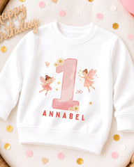 A white sweatshirt with a pink number one and two-tailed fairies printed on it, along with the name 'Annabel' written below.