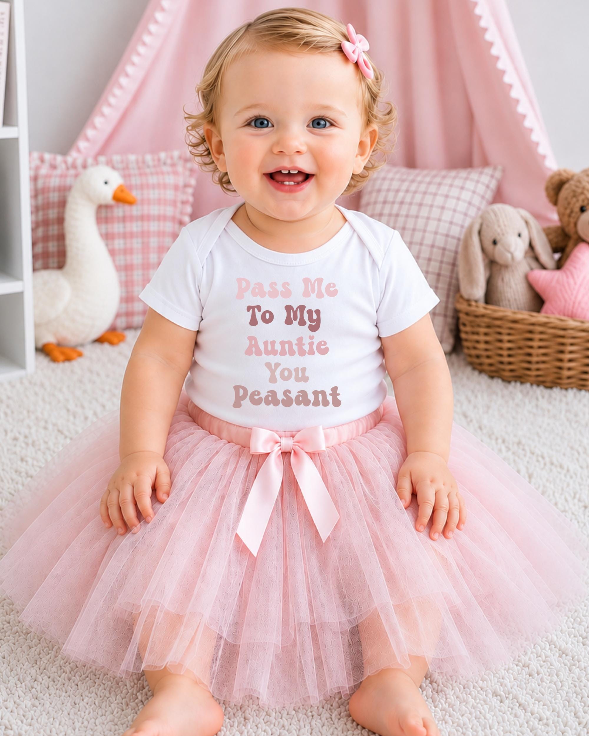 A smiling baby girl wearing a white t-shirt with text and a pink tutu sits on a white carpet, surrounded by stuffed animals and a pink canopy.