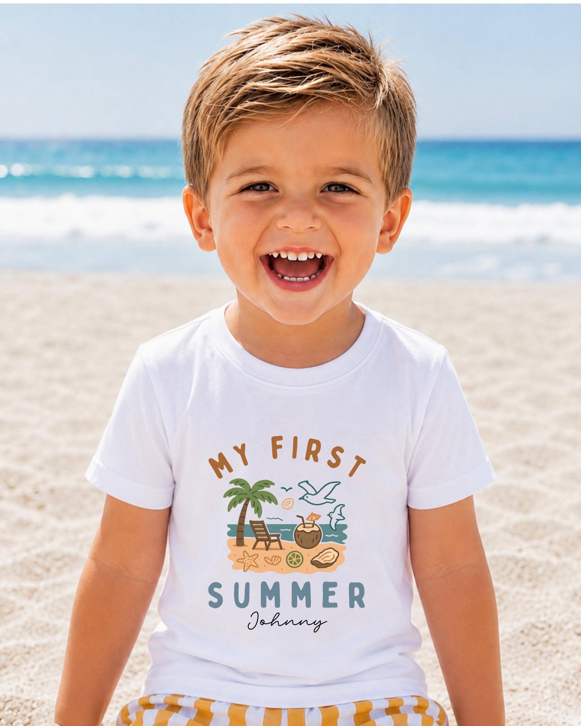 A young boy with a big smile on his face, wearing a white t-shirt with a graphic design of a beach scene and the text "My first summer" printed on it.