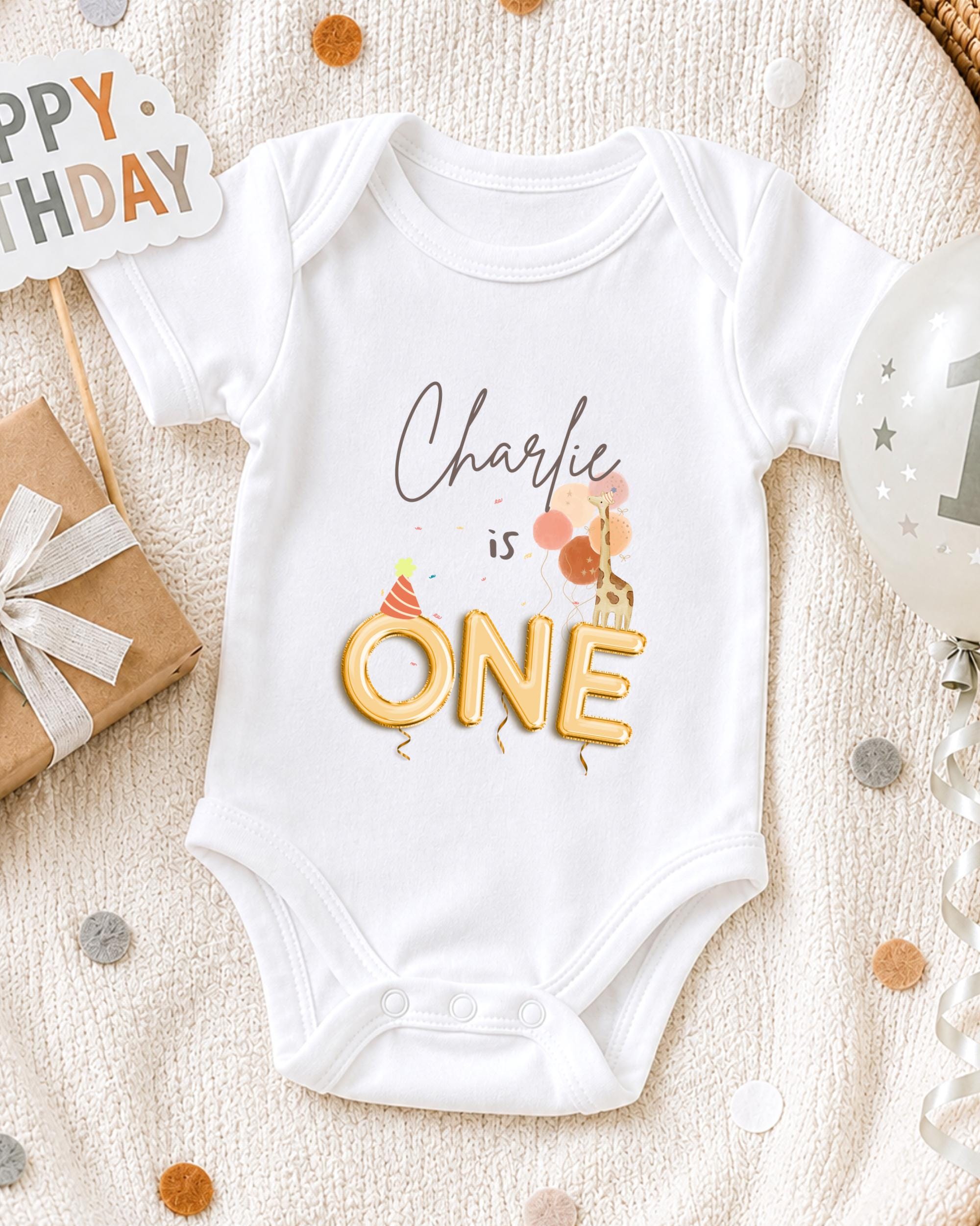 A white onesie with the text "Charlie is ONE" printed on it, along with a birthday balloon and confetti, suggesting a celebration for a baby's first birthday.