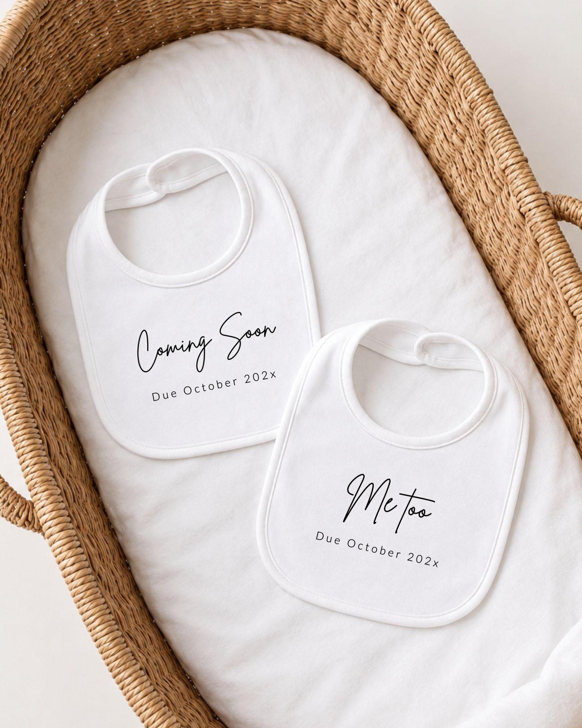 two white bibs with black text, placed in a woven basket. The text on the bibs announces the upcoming arrival of a baby, with the dates due to October 2023.