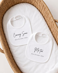 two white bibs with black text, placed in a woven basket. The text on the bibs announces the upcoming arrival of a baby, with the dates due to October 2023.