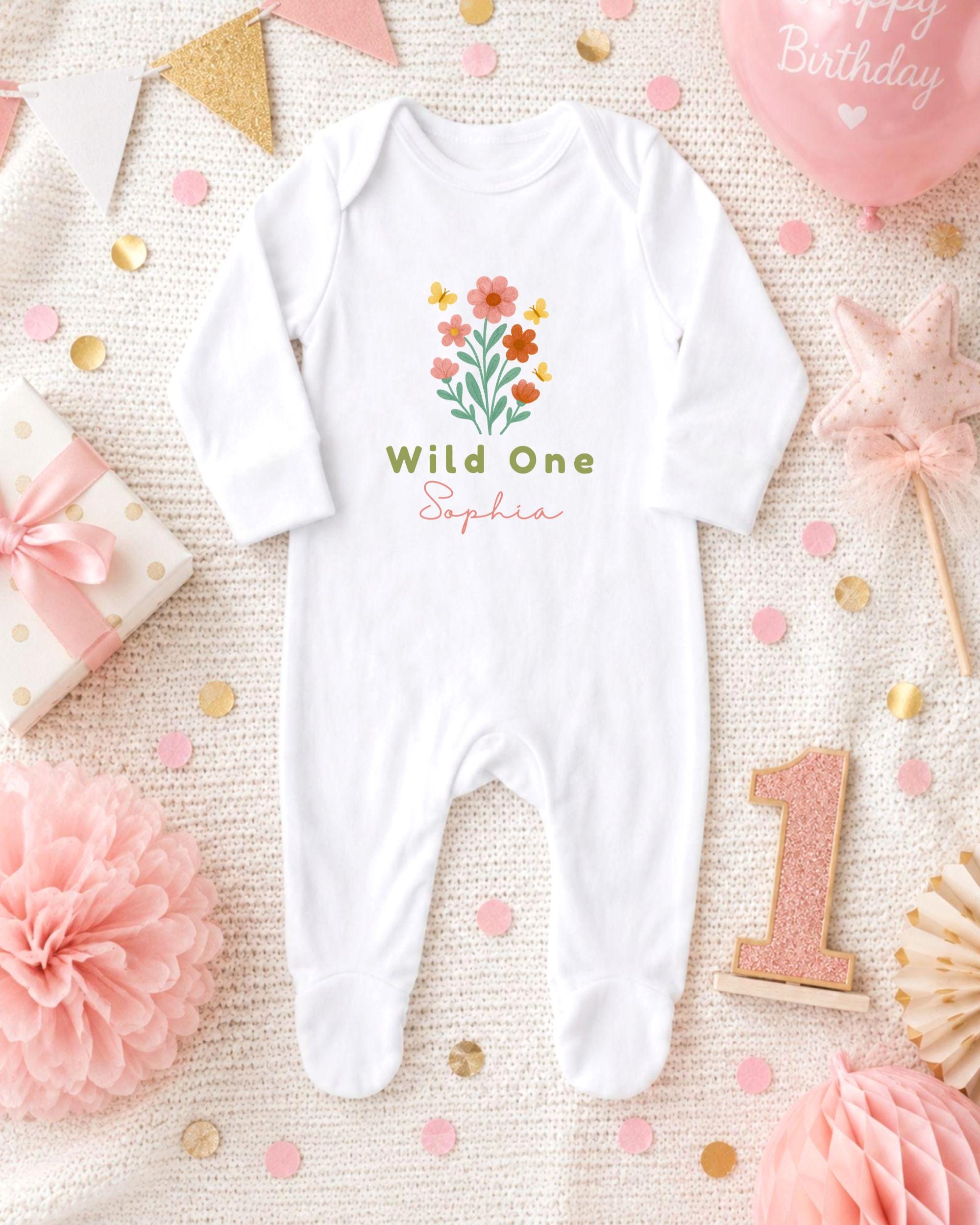 A white onesie with a flower design and the text "Wild One Sophia" printed on it, surrounded by pink and gold confetti, a pink star, and a pink gift box.