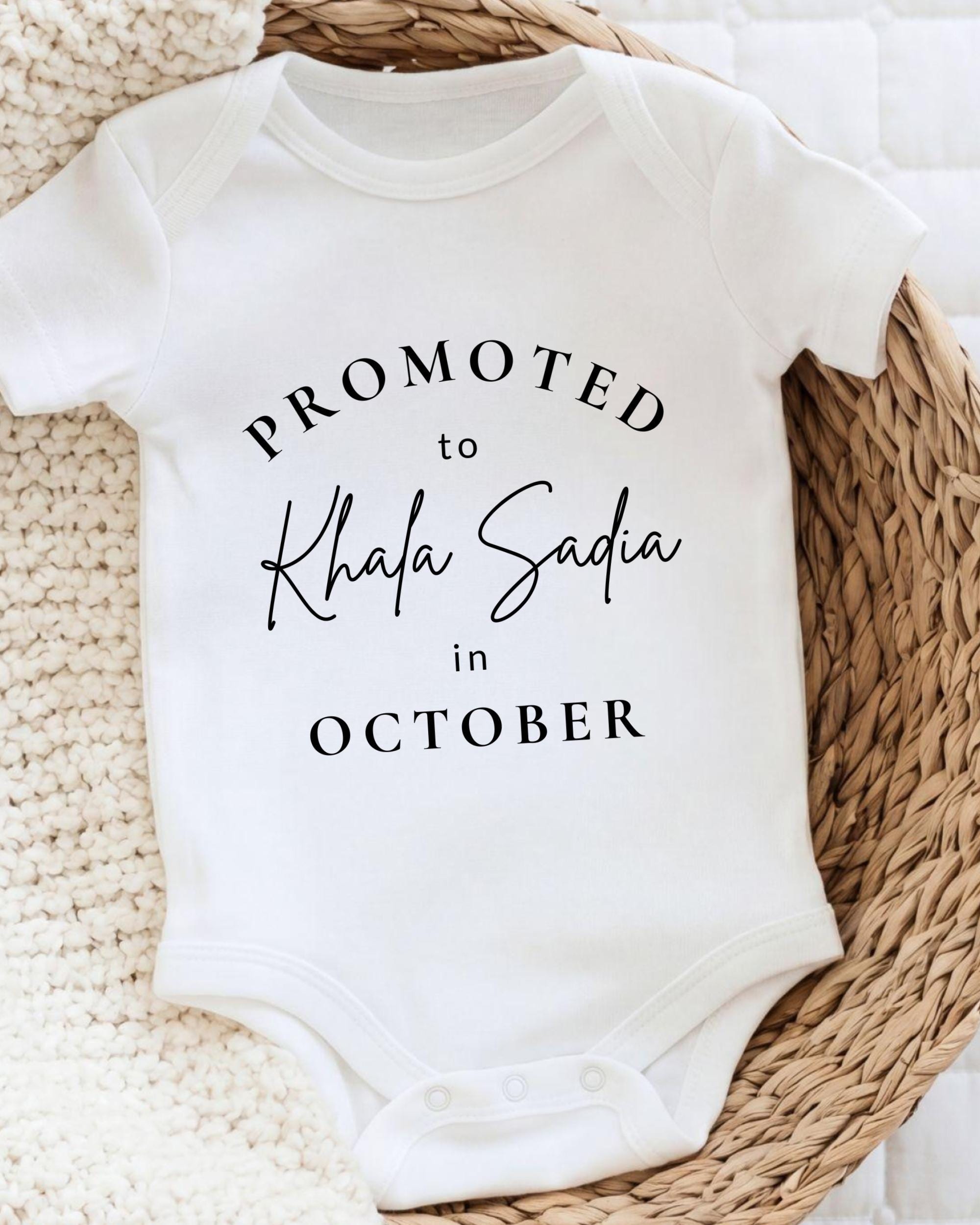 A white baby onesie with the text "PROMOTED to Khala Sadia in October" printed on it.
