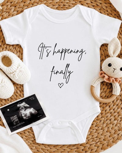White Baby Vest with black script 'It's happening, finally' and an outline heart for a IVF pregnancy announcement.