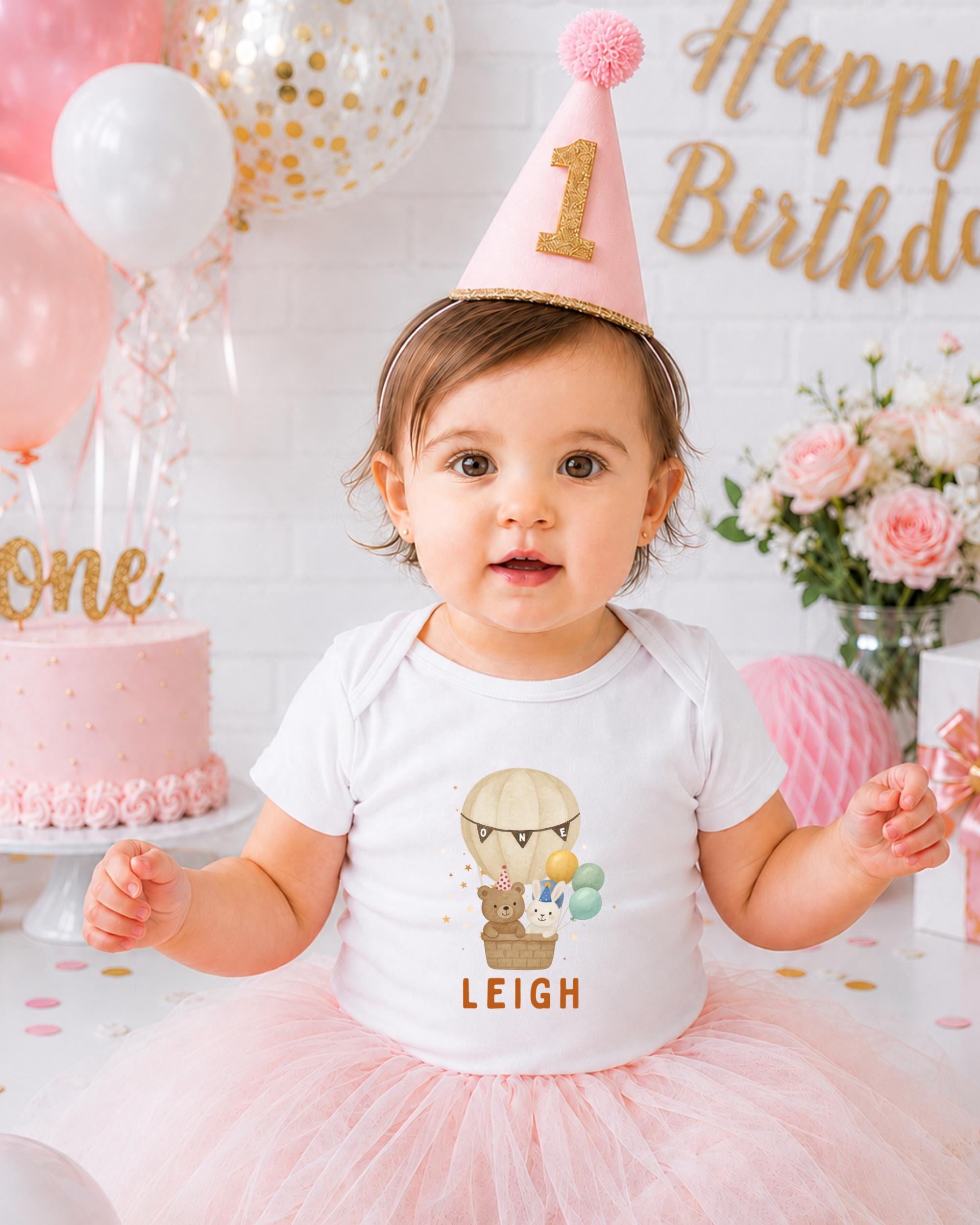 A young girl wearing a birthday hat and a white t-shirt with a cartoon character is posing in front of a birthday cake and balloons.