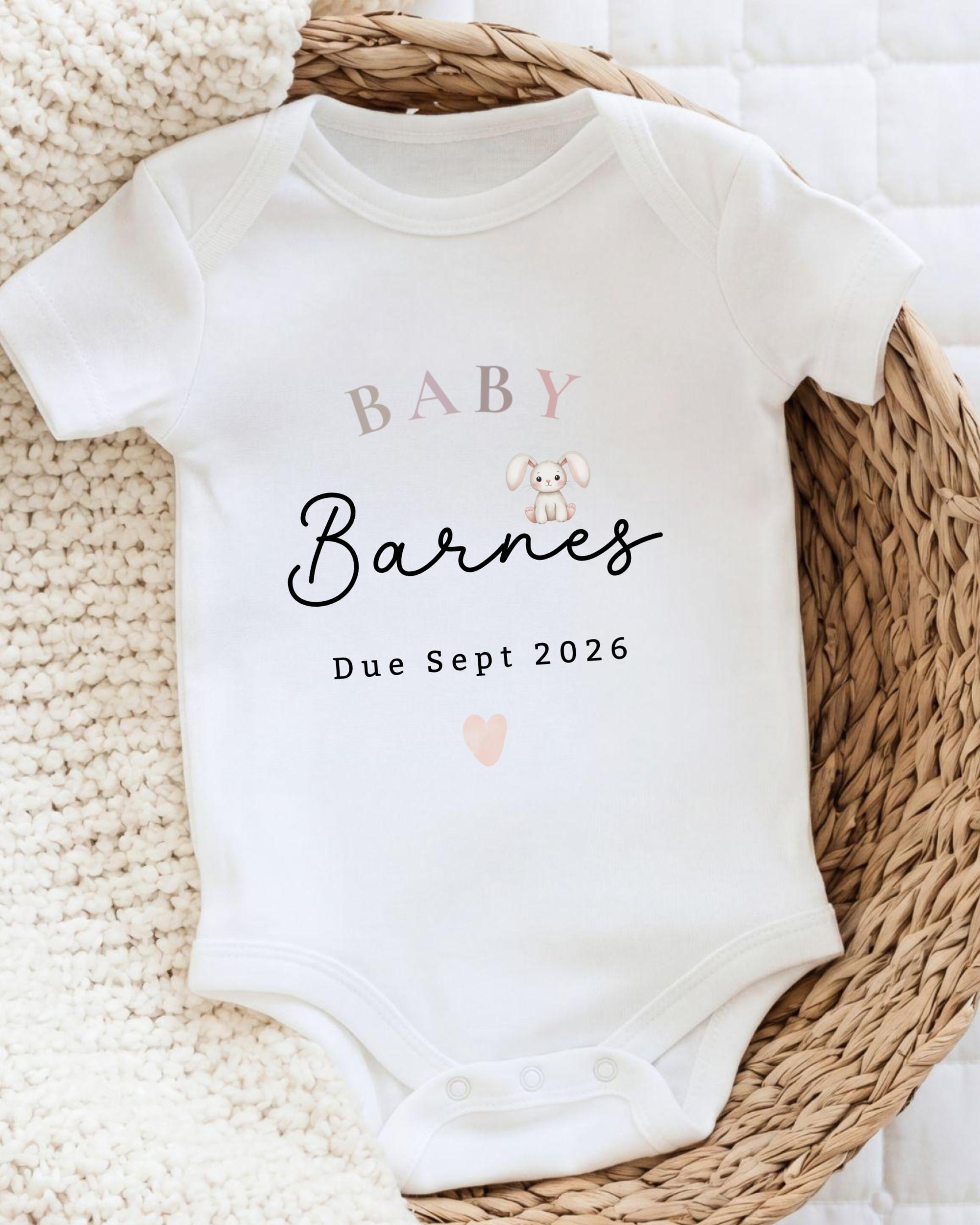 A white baby onesie with the text "BABY Barnes Due Sept 2026" printed on it, placed on a woven basket.