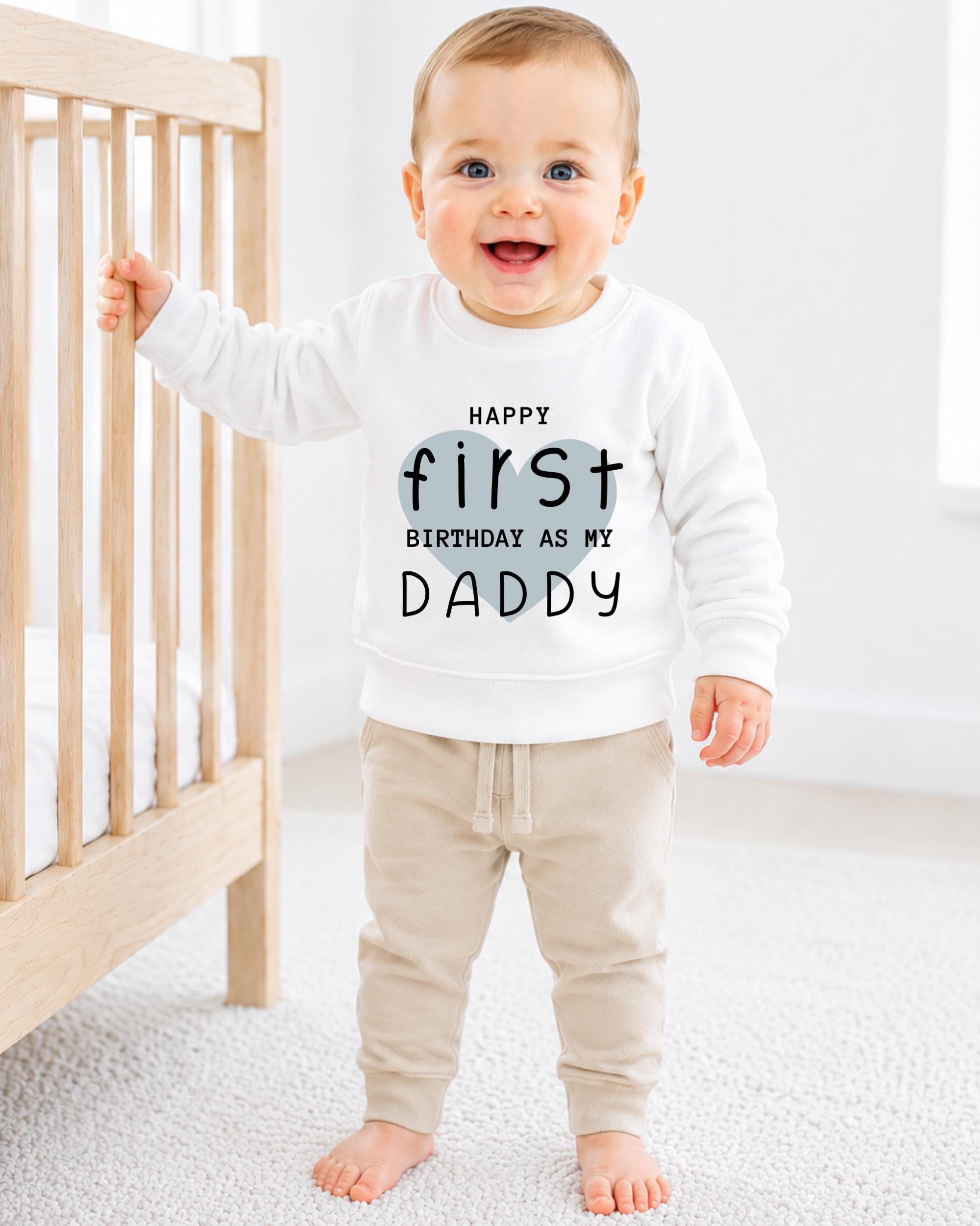 A smiling baby boy wearing a white sweatshirt with text that says "Happy first birthday as my Daddy".