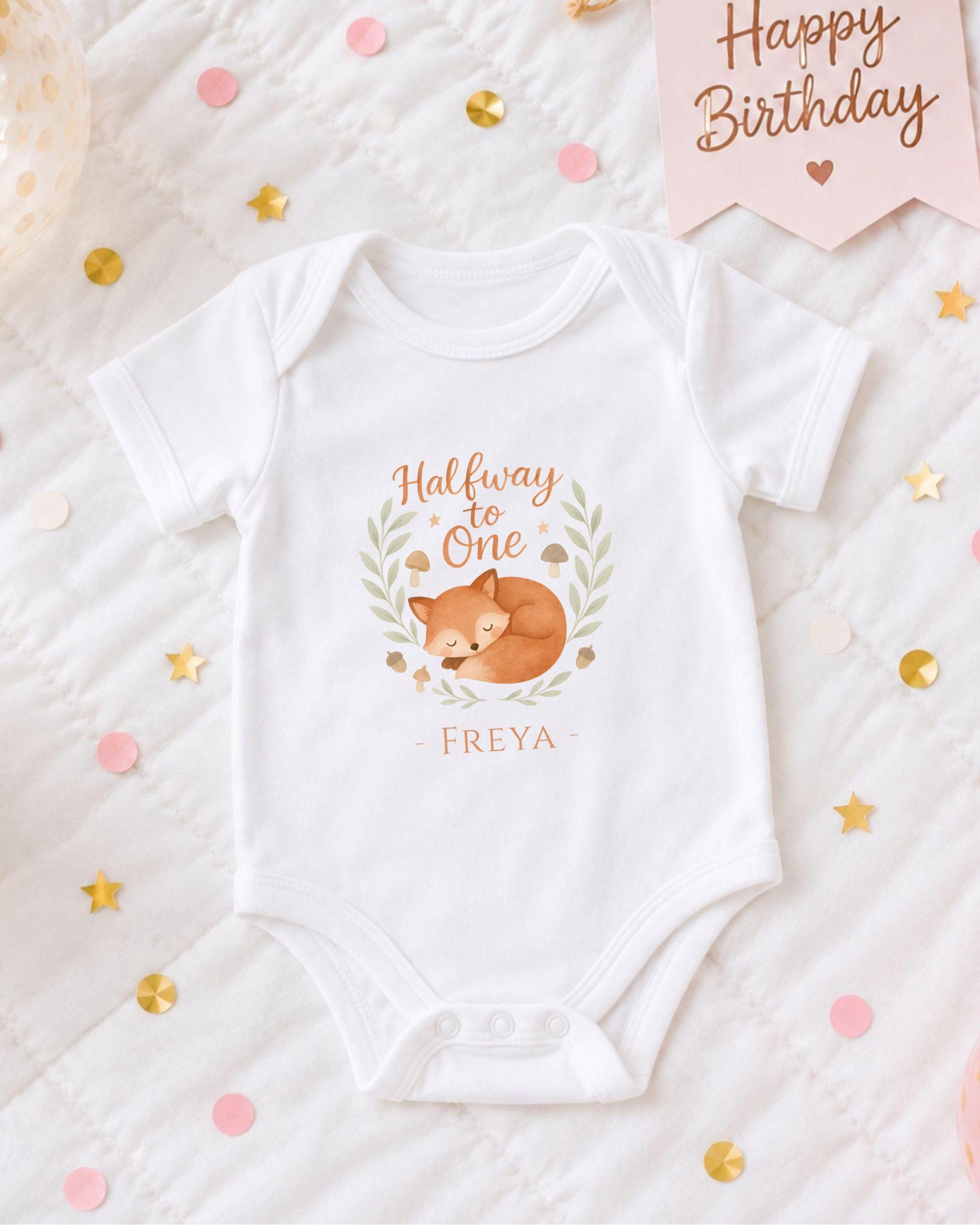 A white onesie with a cartoon fox design and the text "Halfway to One" printed on it, placed on a bed with confetti and a "Happy Birthday" sign.