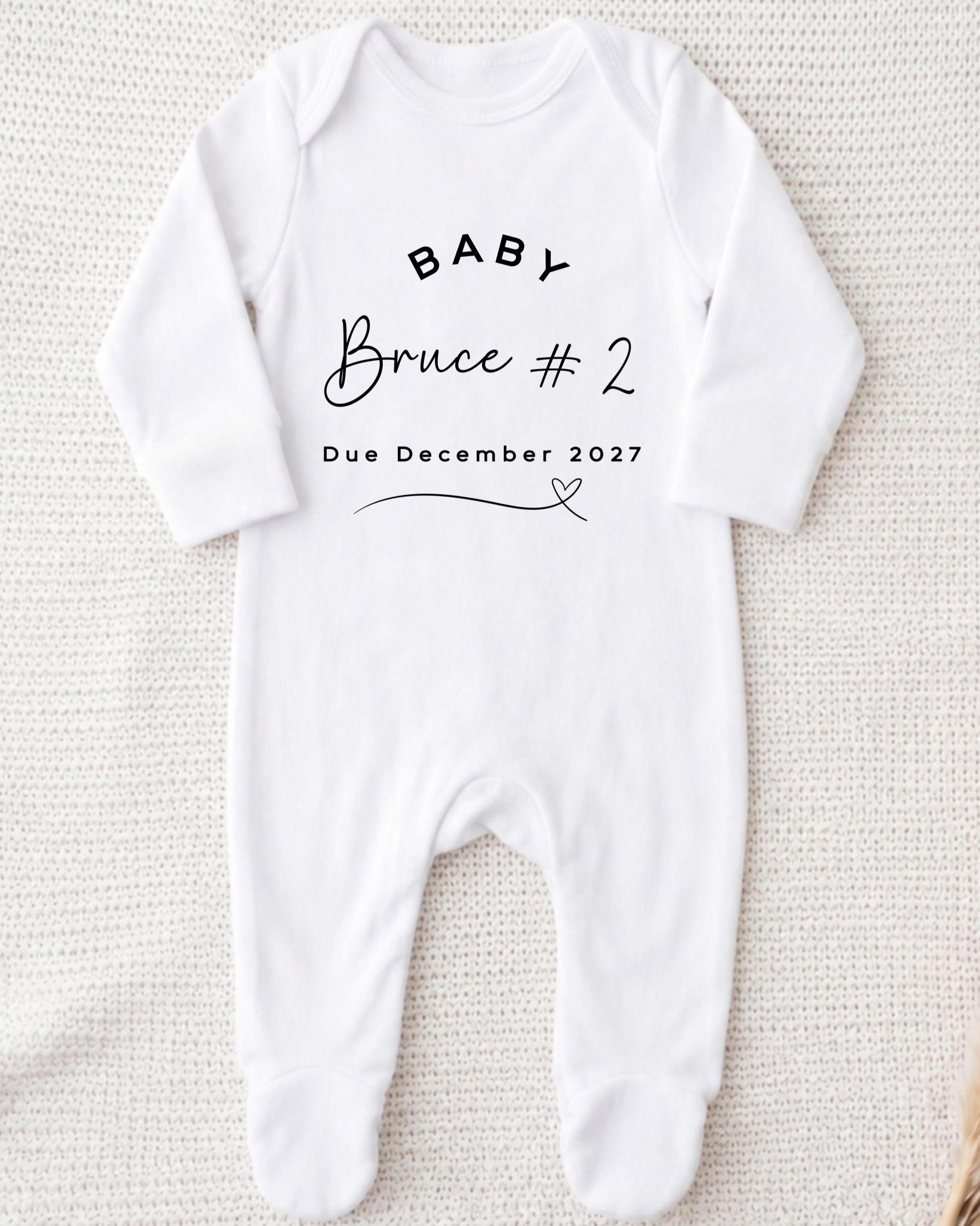 A white onesie with the text "BABY Bruce #2 Due December 2027" printed on it.