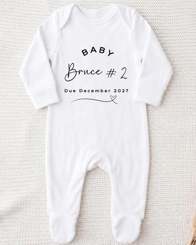 A white bodysuit with the text "BABY Bruce #2 Due December 2027" printed on it.